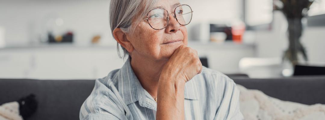 Sad elderly woman sits alone on a sofa at home, looking out the window with a worried expression. She reflects on challenges and feels stressed, illustrating loneliness and concern.