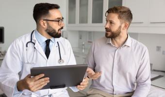 Hispanischer männlicher Arzt, der dem Patienten nach der Untersuchung Beratung gibt, die Ergebnisse der Untersuchung erklärt, Diagnose, auf der medizinischen Couch sitzend, mit dem Mann spricht, medizinische Unterlagen verwendet, Kissen mit Akte hält