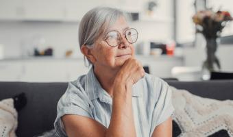 Sad elderly woman sits alone on a sofa at home, looking out the window with a worried expression. She reflects on challenges and feels stressed, illustrating loneliness and concern.
