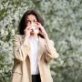 Woman allergic suffering from seasonal allergy at spring in blossoming garden at springtime. Young woman sneezing and blowing nose with nasal handkerchief in front of blooming tree. Allergy concept.